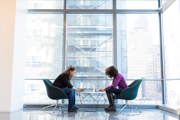 Two women sit facing each other on padded chairs, backlit by floor-to-ceiling windows, working over something on the low table between them. Article image for "How to Answer the Most Common Interview Questions (with Examples)"