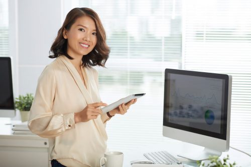 Portrait of pretty Vietnamese businesswoman posing with digital tablet near computer with statistical data on screen