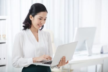 Beautiful young Asian woman in elegant outfit holding and using laptop with smile in modern office