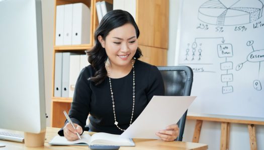 Smiling Asian female entrepreneur reading document and taking notes in planner