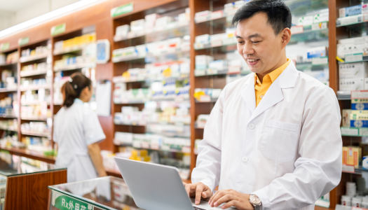 male pharmacist typing on a laptop with medicines in the background