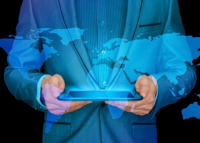 Businessman in a blue suit holding a tablet. A translucent blue electronic visual of the world map is layered on top of the man's torso. Feature image for "Develop These Skills to Compete in Today's Global Job Market"